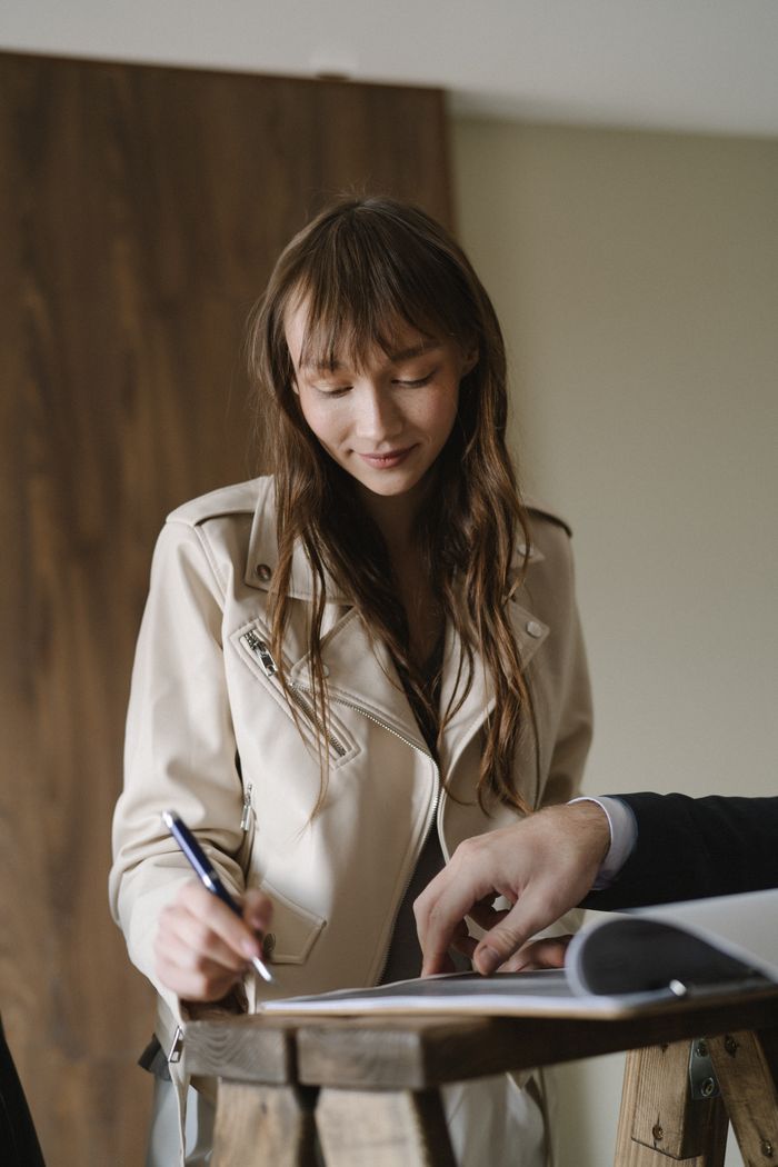 A young woman signing a business contract at an indoor table, holding a pen with focus and intent.